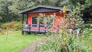 A red cabin with a porch, surrounded by lush greenery and trees. - Lodge 1 "ash" (Isle of Arran)