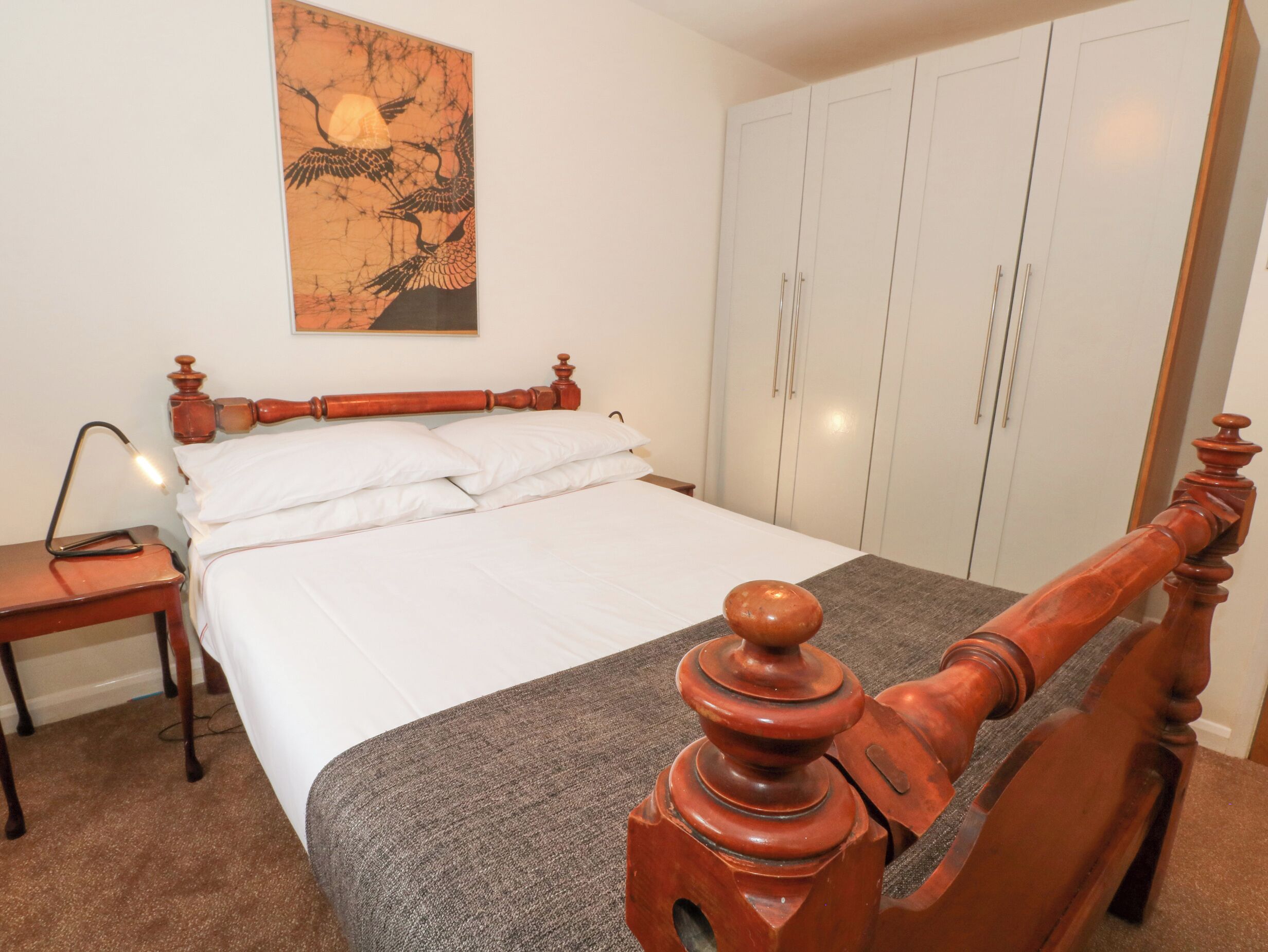 A bedroom with a wooden bed, white bedding, a nightstand, a lamp, and a wardrobe.