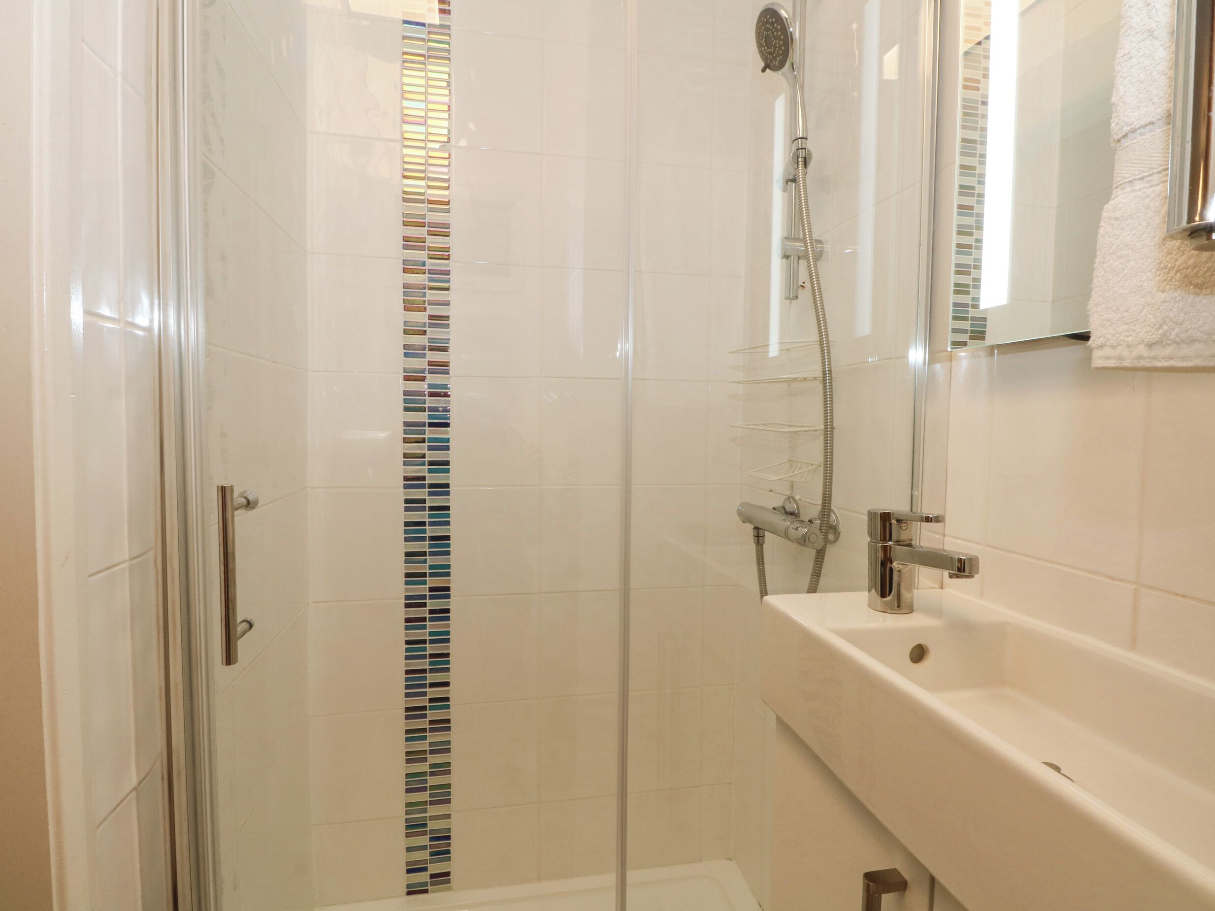 A glass-enclosed shower with a chrome showerhead and handheld shower attachment, a white tiled wall with a vertical mosaic strip, and a white sink with a mirror above it.