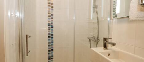 A glass-enclosed shower with a chrome showerhead and handheld shower attachment, a white tiled wall with a vertical mosaic strip, and a white sink with a mirror above it.