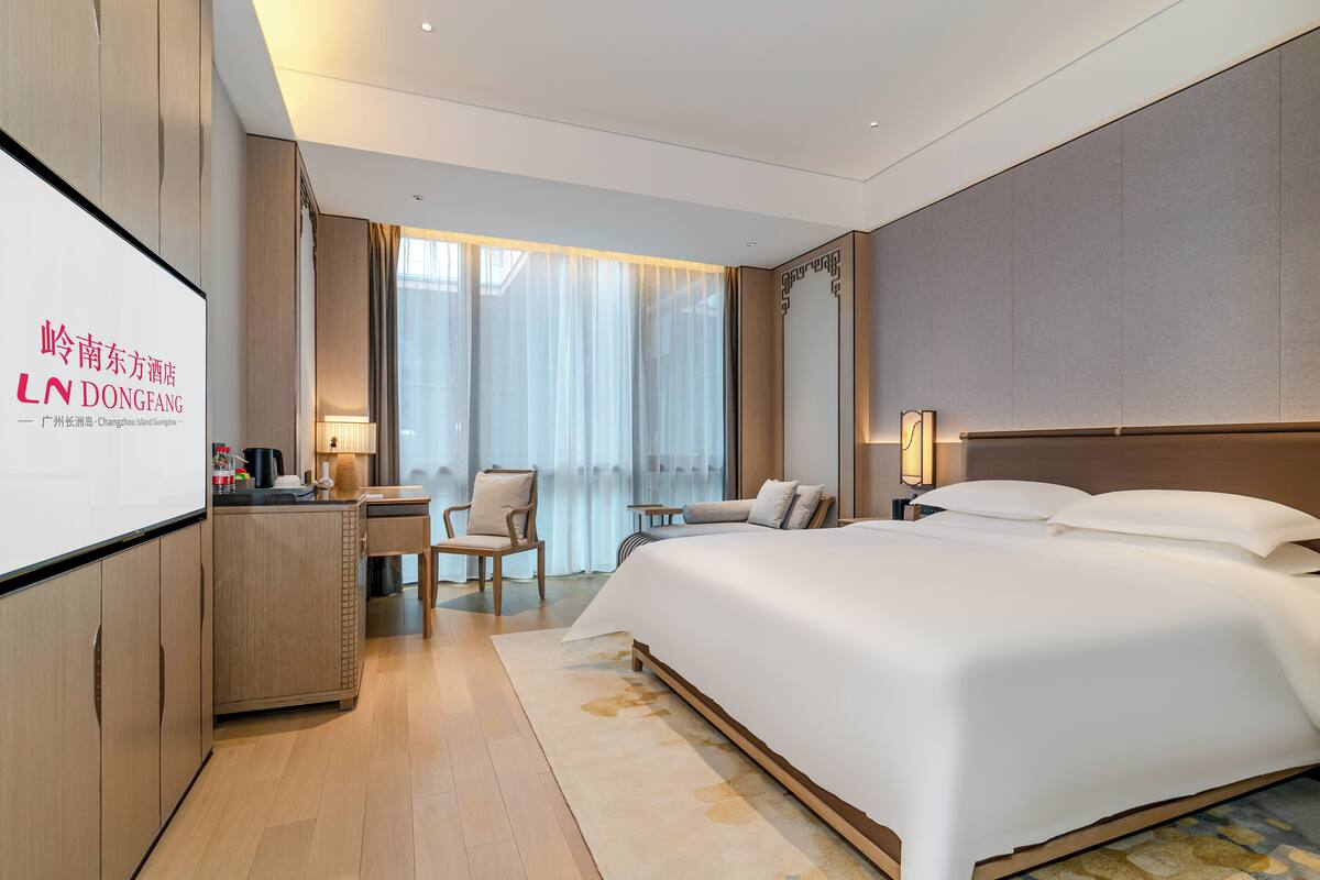 A hotel room with a large bed, a desk with a chair, a TV screen, and a view of the outdoors through large windows.