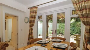 A dining area with a wooden table set for four, a clock on the wall, and a view of a garden through glass doors. - The Coach House Apartment (Dawlish)