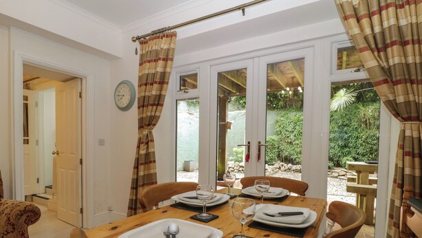A dining area with a wooden table set for four, a clock on the wall, and a view of a garden through glass doors.