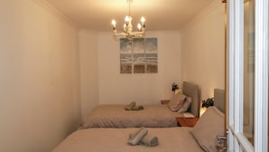 A bedroom with two beds, a chandelier, a painting, and a window with a view.
