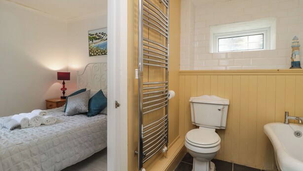 A bedroom with a bed, a bedside table with a lamp, a window, and a bathroom with a toilet and sink. - The Coach House Apartment (Dawlish)