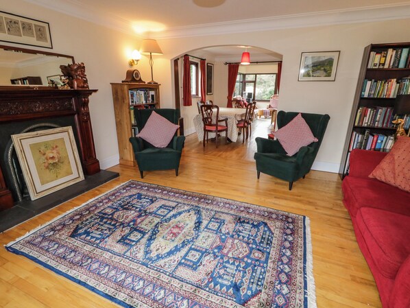 A living room with a wooden floor, green armchairs, a red sofa, a patterned rug, a fireplace, a bookshelf, and a dining area with a table and chairs. - Tudor Lodge (Newcastle)