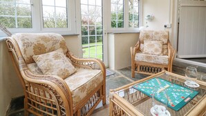 Quarto com duas cadeiras de vime com tecido floral, uma mesa de vidro com um jogo de tabuleiro e grandes janelas com vista para um jardim.