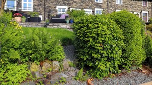 A stone house with multiple windows and a well-maintained garden with various plants and a large bush. - Lavender Cottage (Coniston)