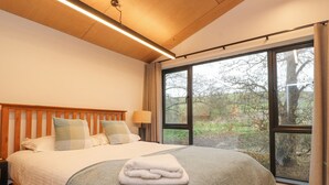 Une chambre à coucher avec un grand lit, une tête de lit en bois et une vue sur les arbres, visible à travers une grande fenêtre.
