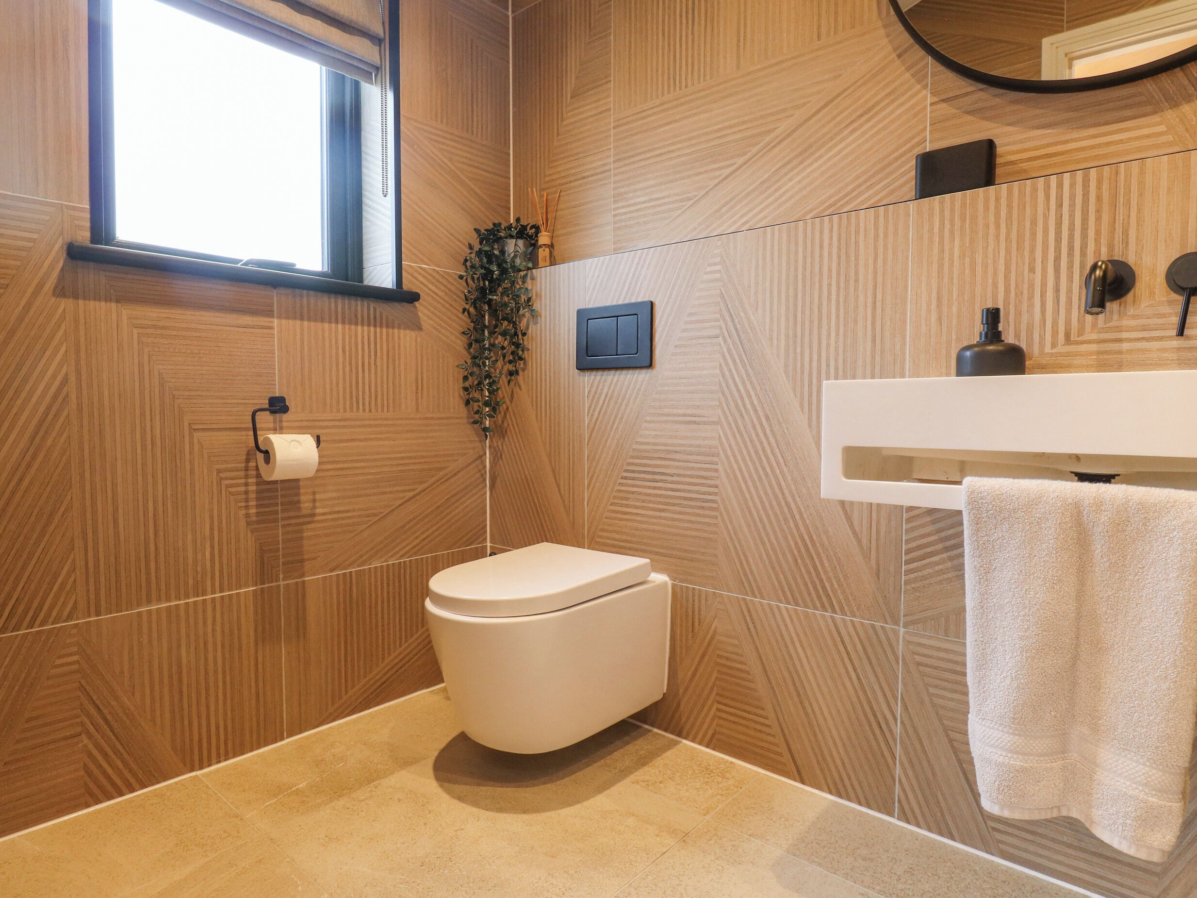 Une salle de bain moderne avec un mur décoré d’un motif en chevrons en bois, des toilettes blanches, un lavabo blanc avec une serviette et un miroir au-dessus du lavabo.