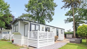 A mobile home with a fenced deck, a small porch, and a garden area with gravel and plants. - Woodlands Lodge (Ruthin)