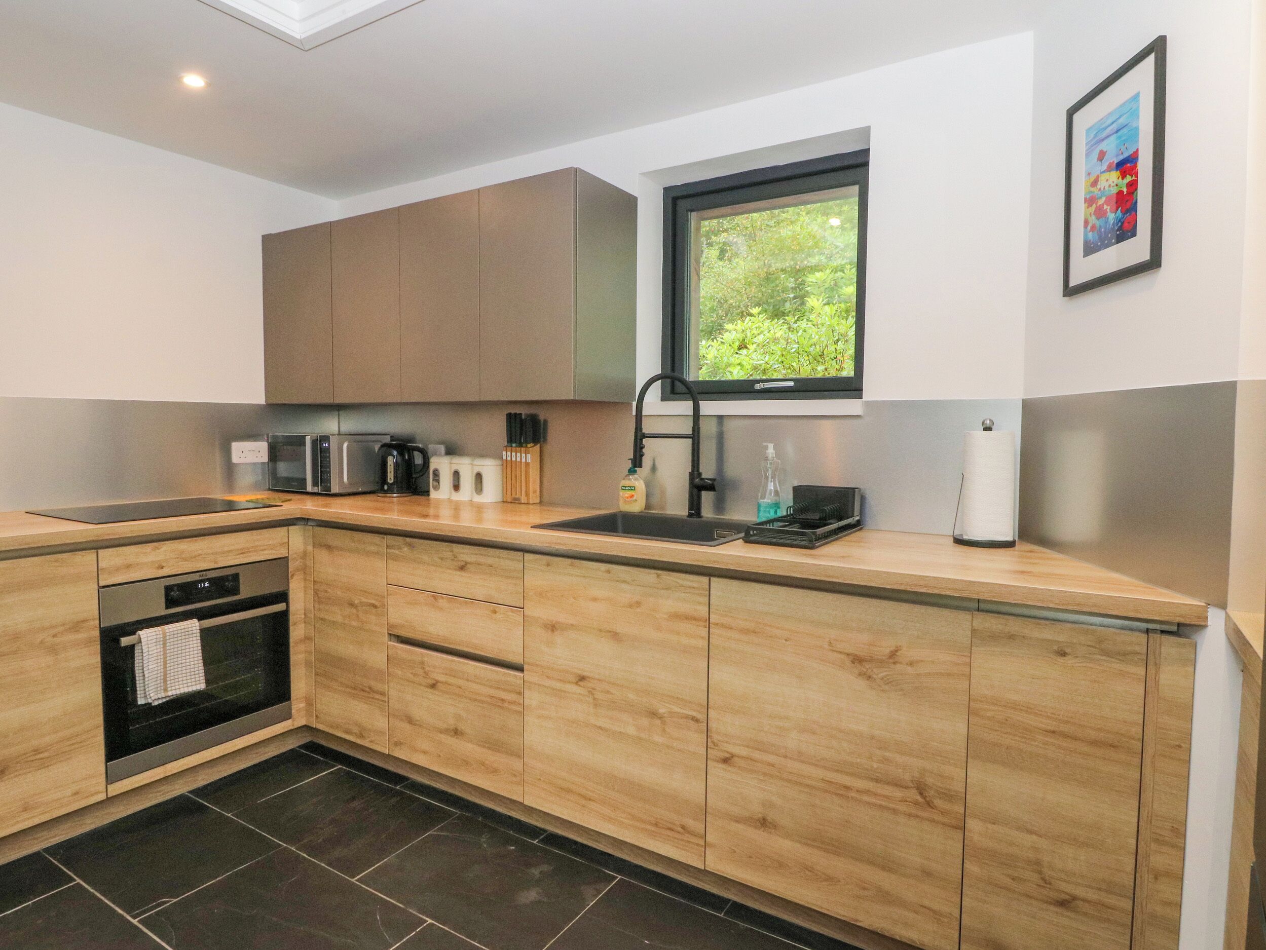 A modern kitchen with wooden cabinets, a built-in oven, a microwave, and a window with a view of greenery.