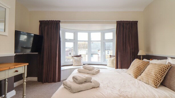 A hotel room with a large bed, a TV on the wall, and a view of the outside through a large window. - Flat 2 Niles Place (Padstow)