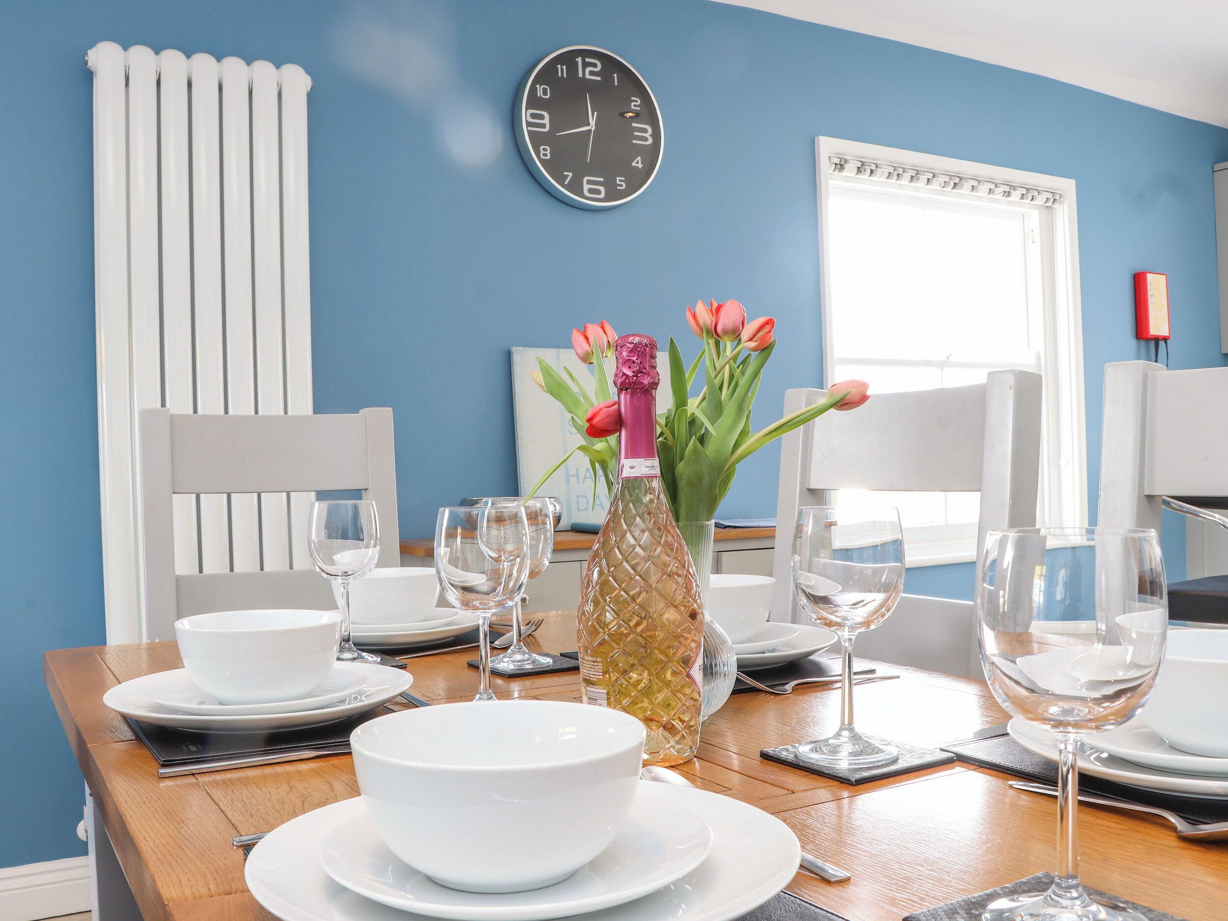 A dining table set with white plates, bowls, and wine glasses, a vase with flowers, and a clock on the wall.