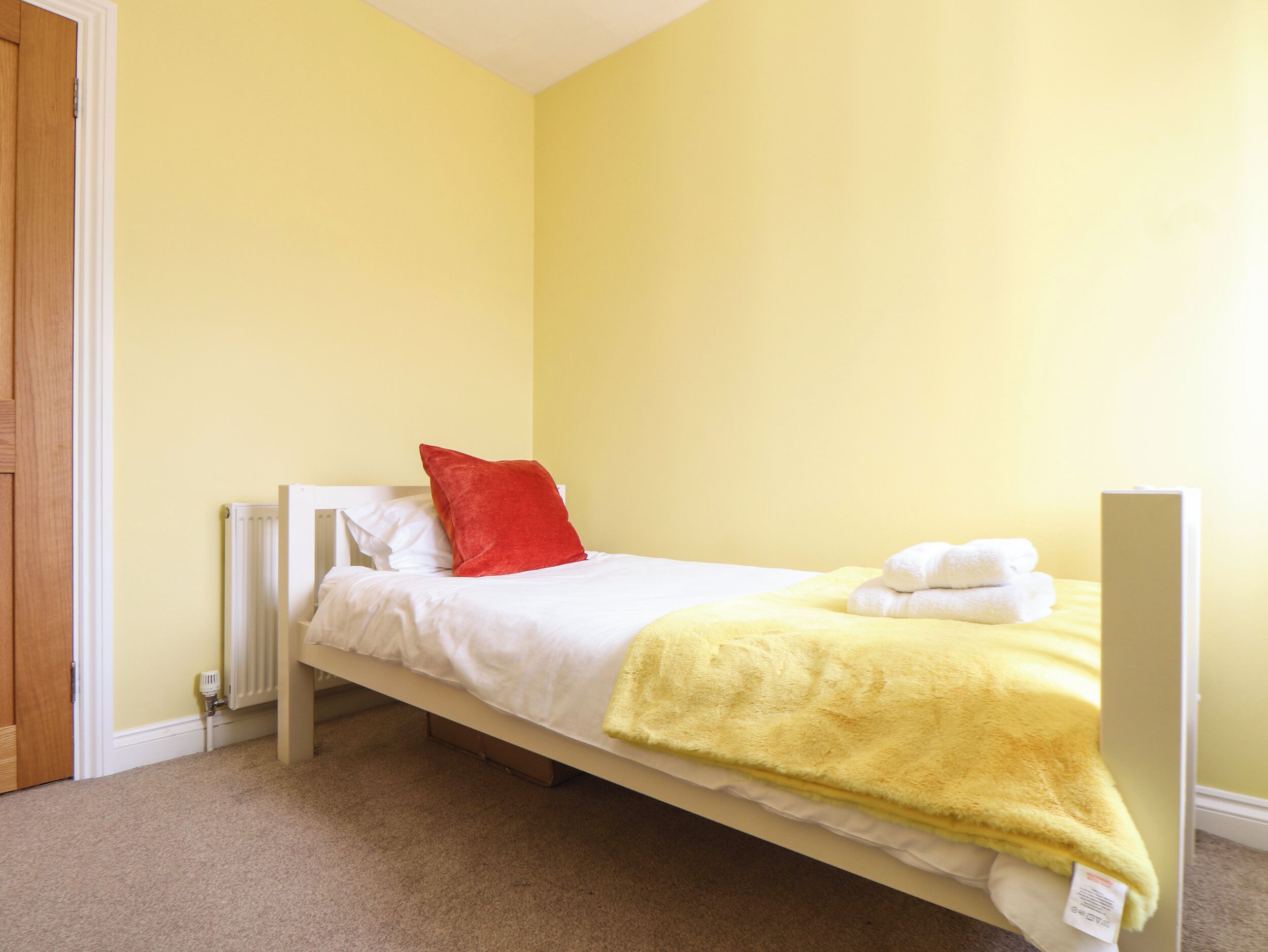 A single bed with white bedding and a red pillow in a room with yellow walls and a wooden door.