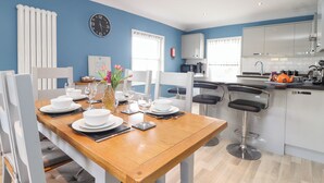 A modern kitchen with white cabinets, a black clock on the wall, and a dining area with a wooden table and white chairs. - Flat 2 Niles Place (Padstow)