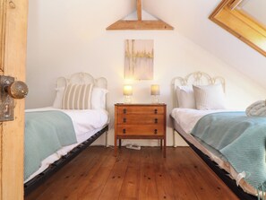 A bedroom with two single beds, a wooden nightstand with drawers, a painting above the nightstand, and a wooden ceiling beam. - Bert's Barn (Helston)