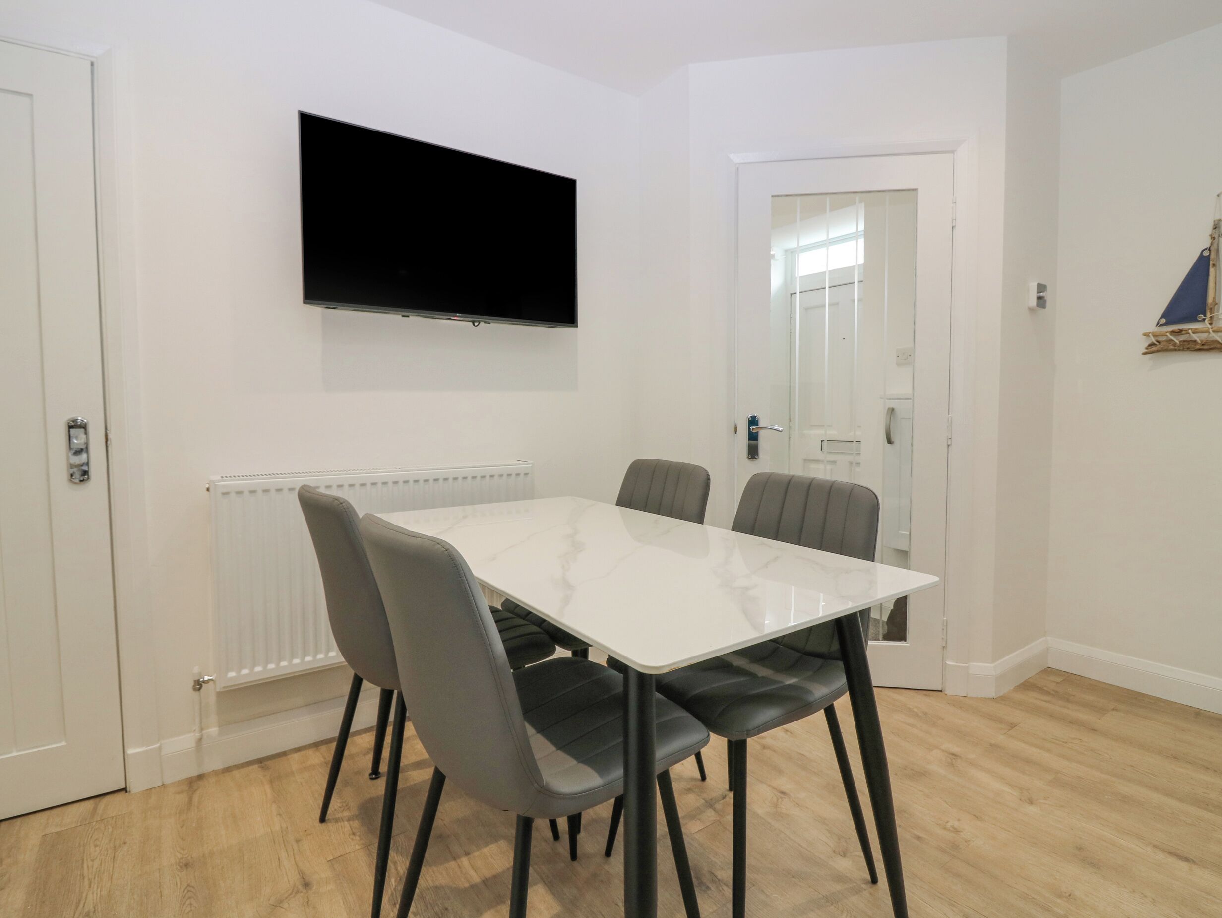 A modern room with a white table and four grey chairs, a flat-screen TV mounted on the wall, and a white door with a mirror.