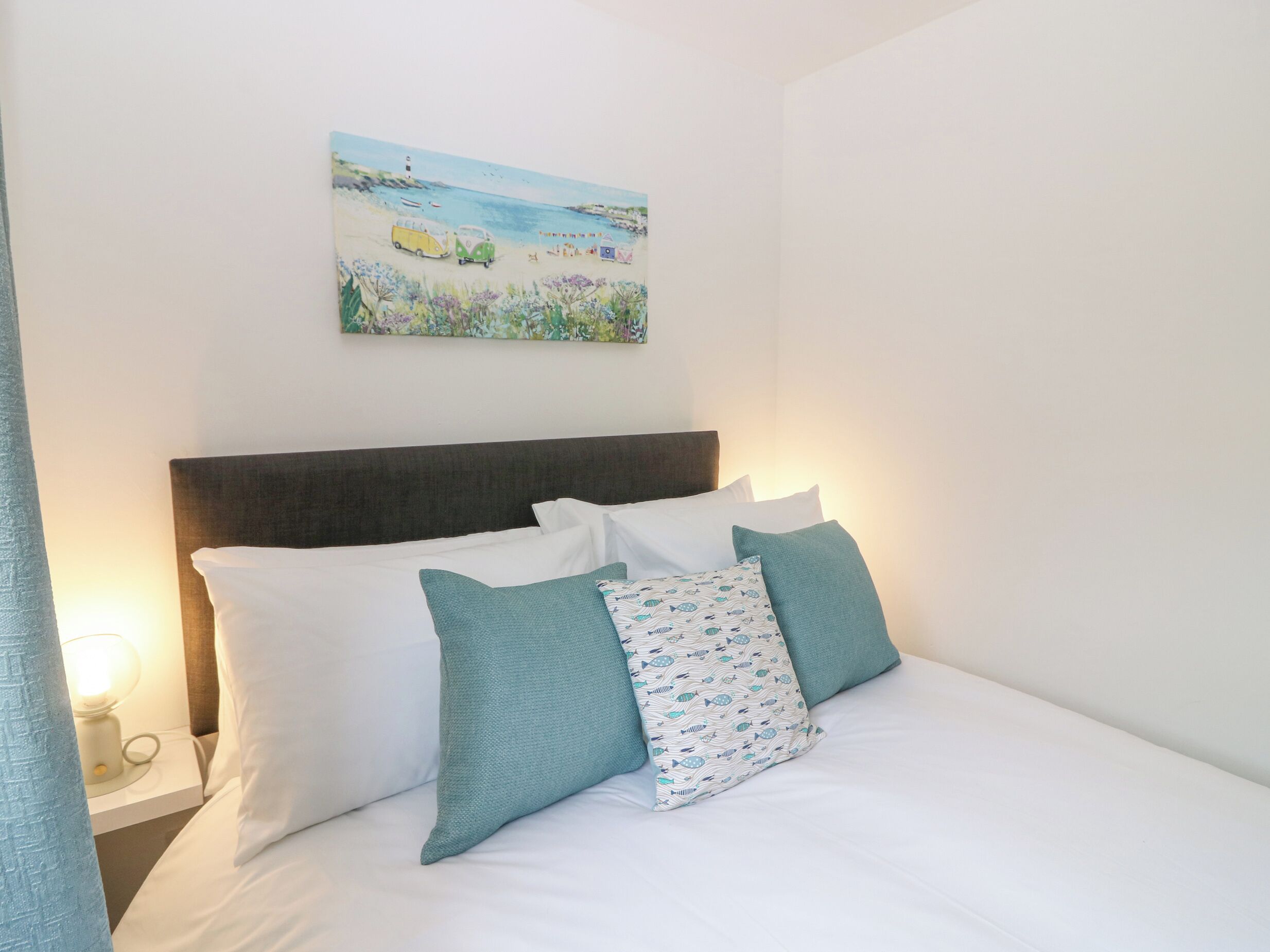 A neatly made bed with white and teal pillows, a dark headboard, and a painting of a beach scene above it.