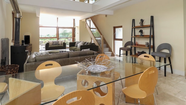 Amplia sala de estar con mesa de vidrio, sillas modernas, un sofá y una escalera que lleva a un nivel superior.