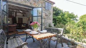 Uma área de refeições ao ar livre com paredes de pedra, uma mesa de madeira posta com pratos e copos, e um banco.
