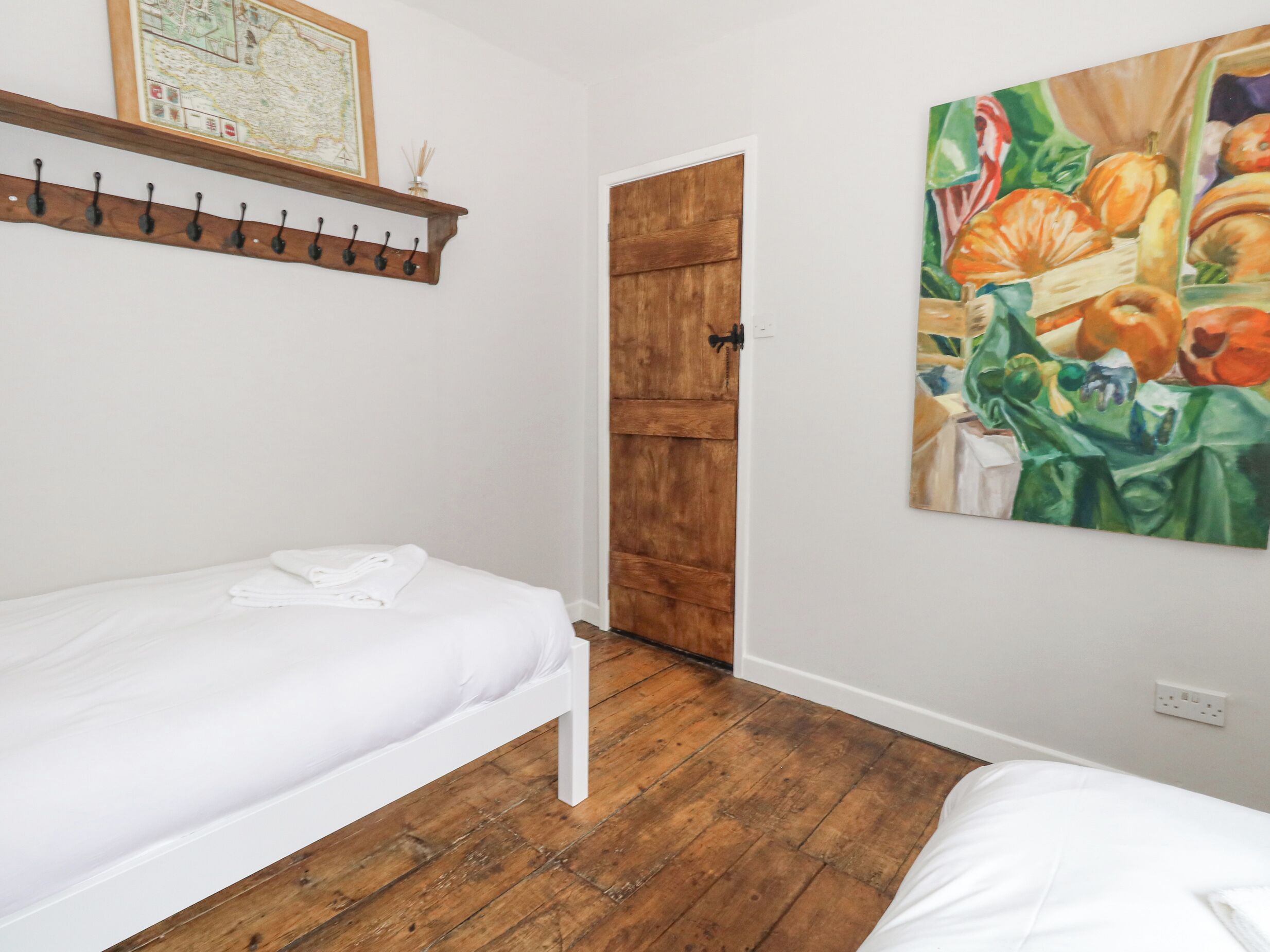 Un dormitorio con suelo de madera, una cama blanca, una puerta de madera y un cuadro de verduras en la pared.