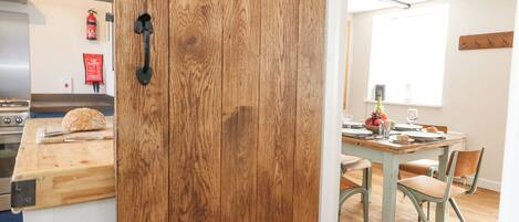 A kitchen with a wooden door, a cutting board with a loaf of bread, and a dining area with a table set for two.