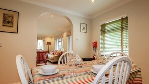 A dining area with a striped tablecloth, white chairs, and a view into a living room with a sofa and a lamp. - 6 Trelawney Court (Wadebridge)