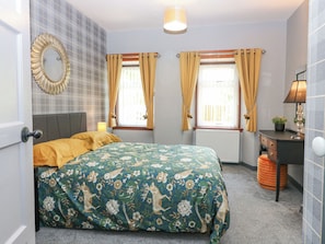 A bedroom with a bed, a bedside table, a lamp, a window with curtains, and a mirror on the wall. - The Cottage (Kirriemuir)