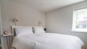 Ein ordentlich bezogenes Bett in einem Zimmer mit weißer Wand, einem kleinen Nachttisch mit Lampe und einem Fenster mit Jalousien.