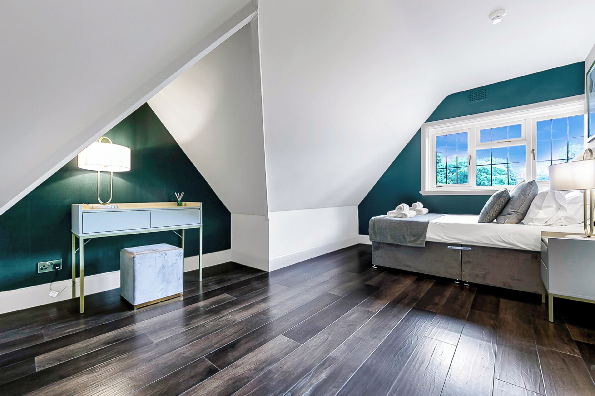 A modern loft bedroom with a sloped ceiling, a desk with a lamp, a bed with pillows, and a window with a view.