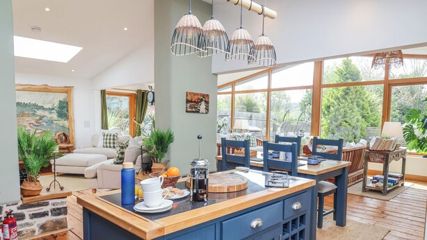 Une cuisine moderne avec un îlot central bleu, un plancher en bois et de grandes fenêtres donnant sur un paysage verdoyant.