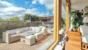 Une terrasse dotée de meubles en osier, d’une table en verre et offrant une vue sur la cuisine et le jardin.