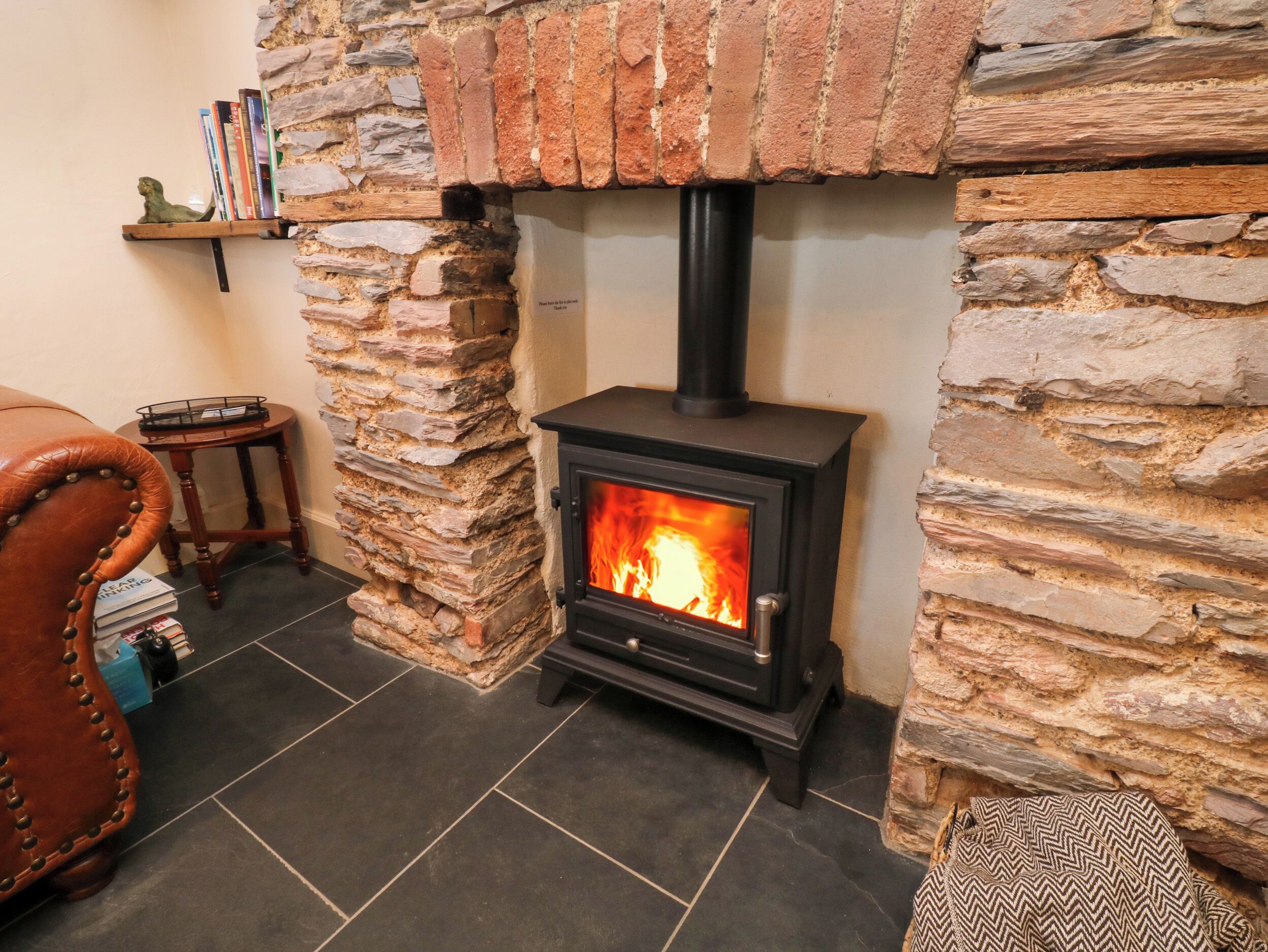 A stone fireplace with a black stove, a leather chair, and a small wooden table.