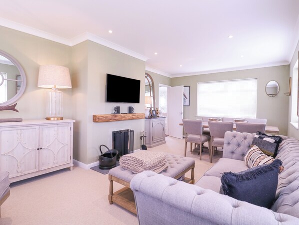A spacious living room with a grey sofa, a coffee table, and a fireplace. There is a dining area with a table and chairs, and a TV mounted on the wall. - The Farmhouse (Clacton-on-Sea)