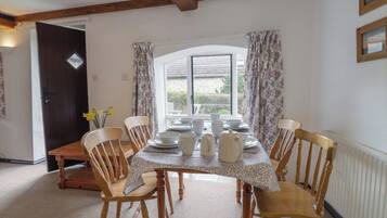 A dining room with a wooden table set for six, surrounded by six wooden chairs. There is a window with floral curtains, a door, and a radiator on the wall.