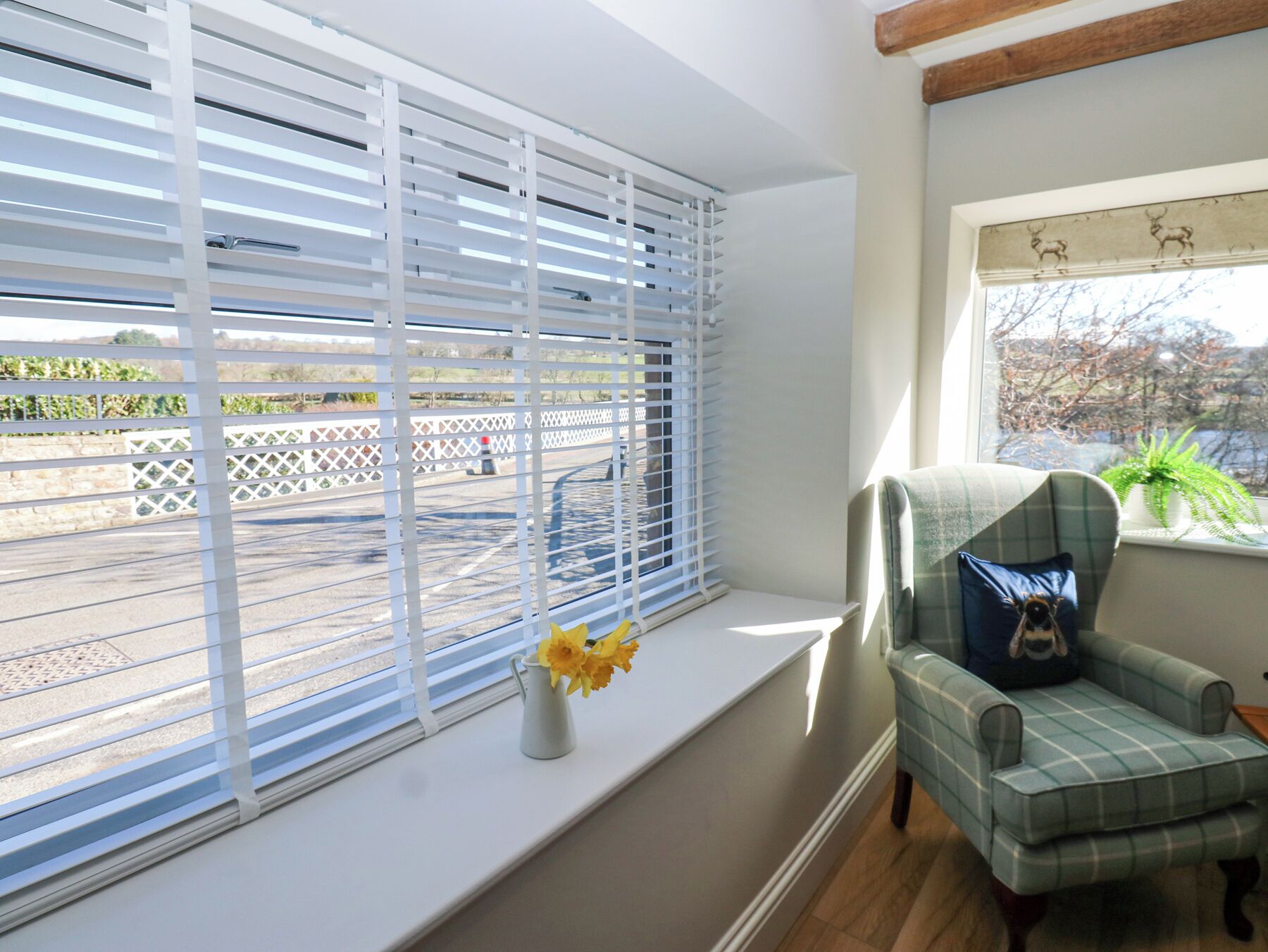 A room with a window with white blinds, a small vase with yellow flowers on the windowsill, and a plaid armchair with a blue pillow.