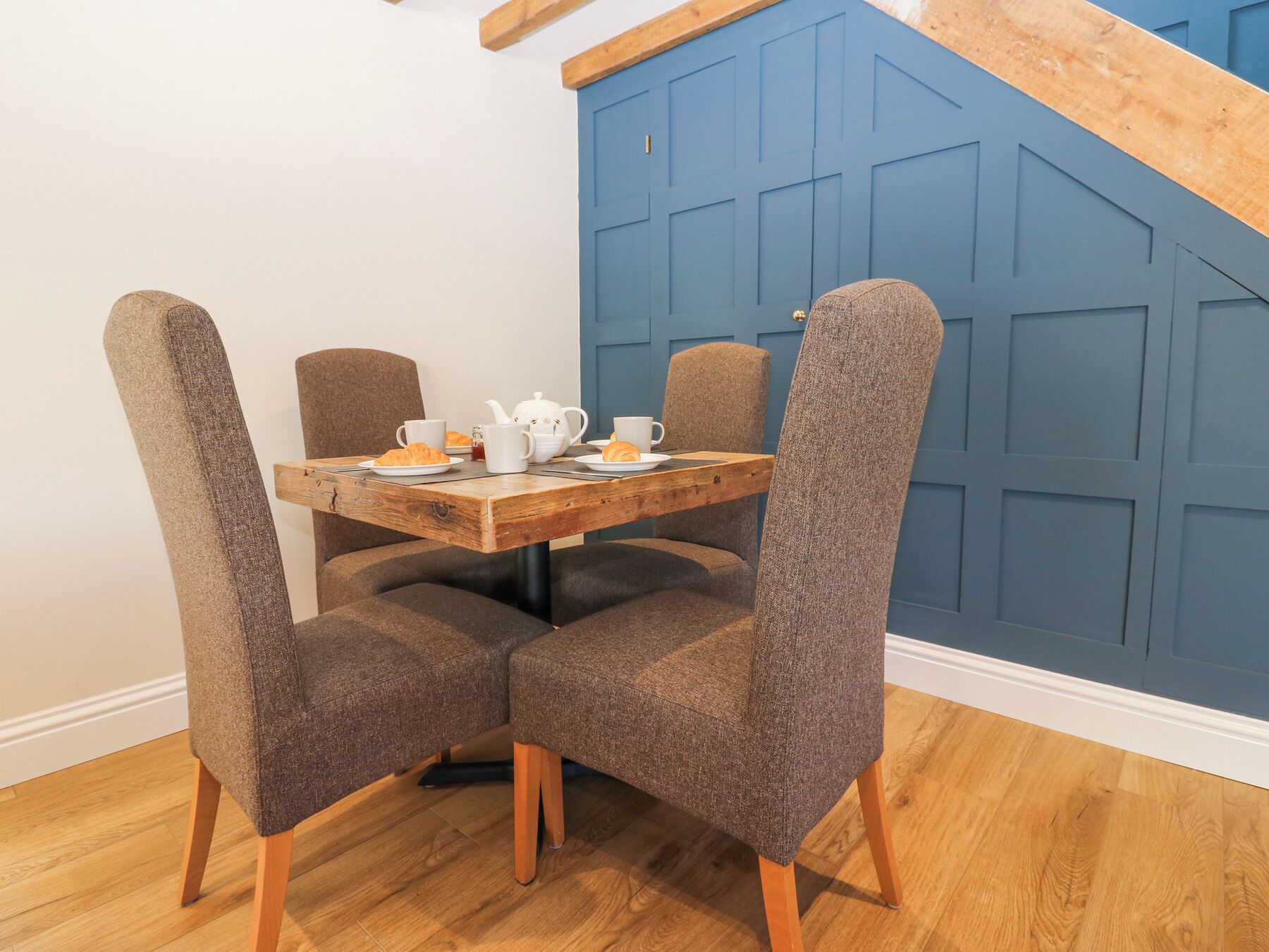 Een eethoek met een houten tafel, vier grijze bekleden stoelen en een blauwe wand met een geometrisch patroon.