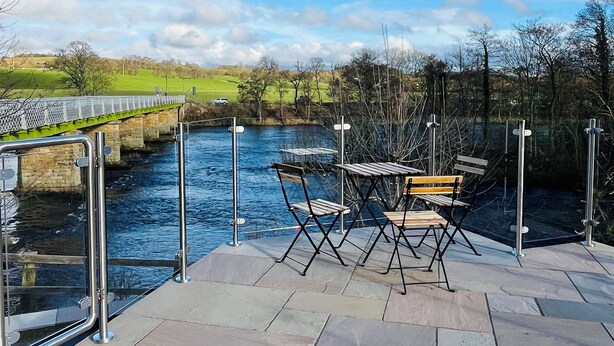 Una terraza junto al río con una mesa y sillas, una barandilla de metal y un puente al fondo.