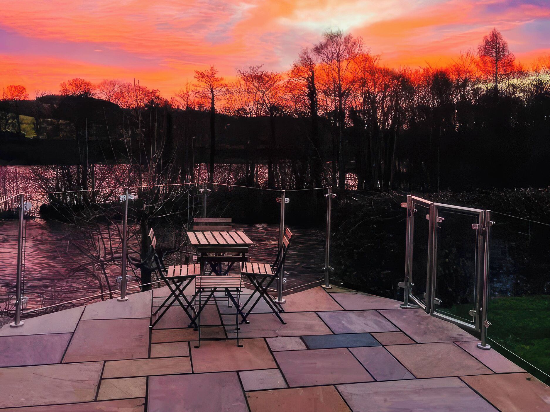 Eine Terrasse mit Tisch und Stühlen, einer Glasreling und Blick auf Bäume sowie ein Gewässer.