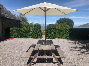 A wooden picnic table with a large white umbrella, set on a gravel surface with a stone building and green hedges in the background. - Worthy Barn (Minehead)