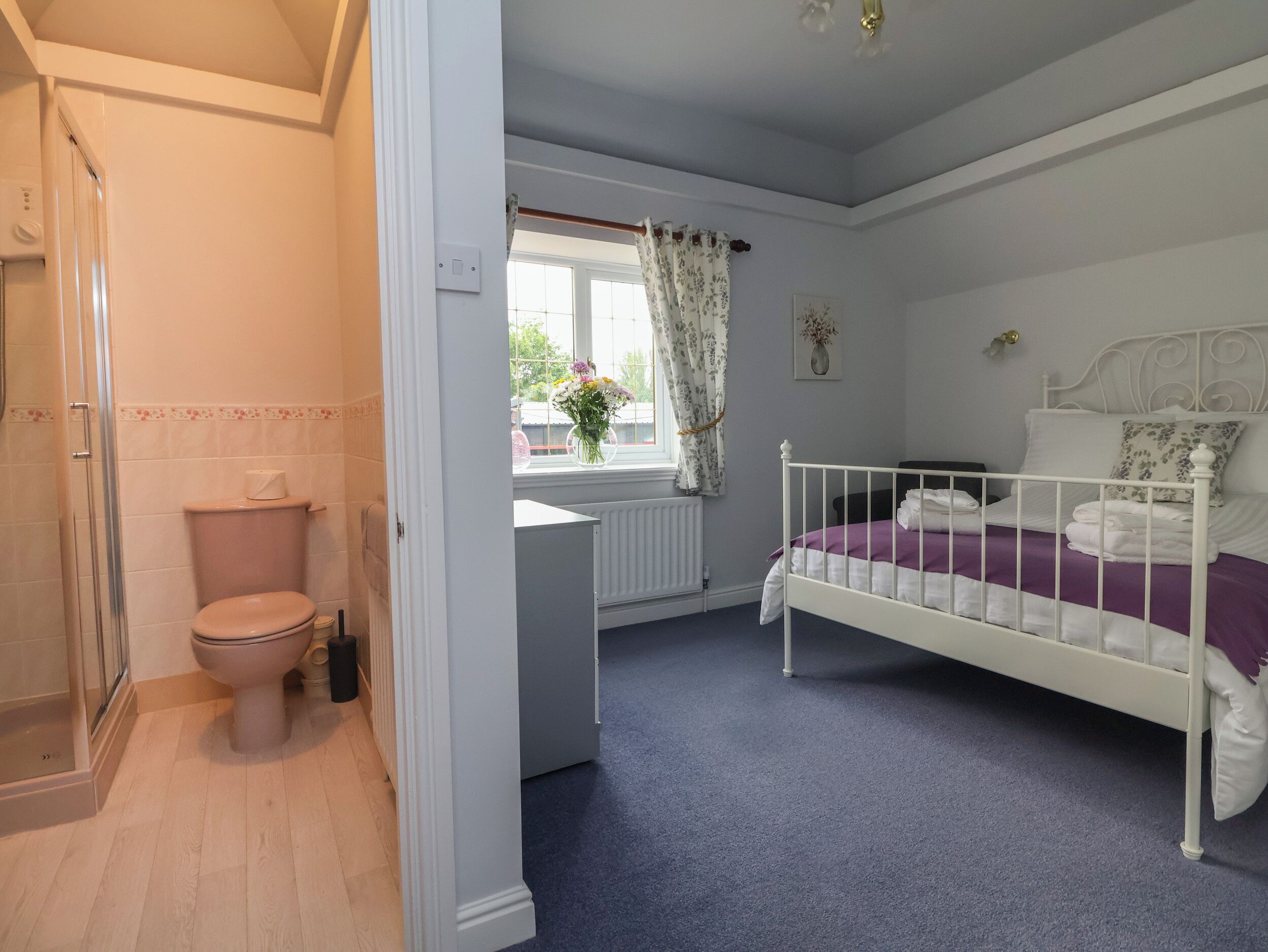 Une chambre à coucher avec un lit blanc, une couvre-lit violette et une fenêtre ornée de rideaux à motifs floraux.