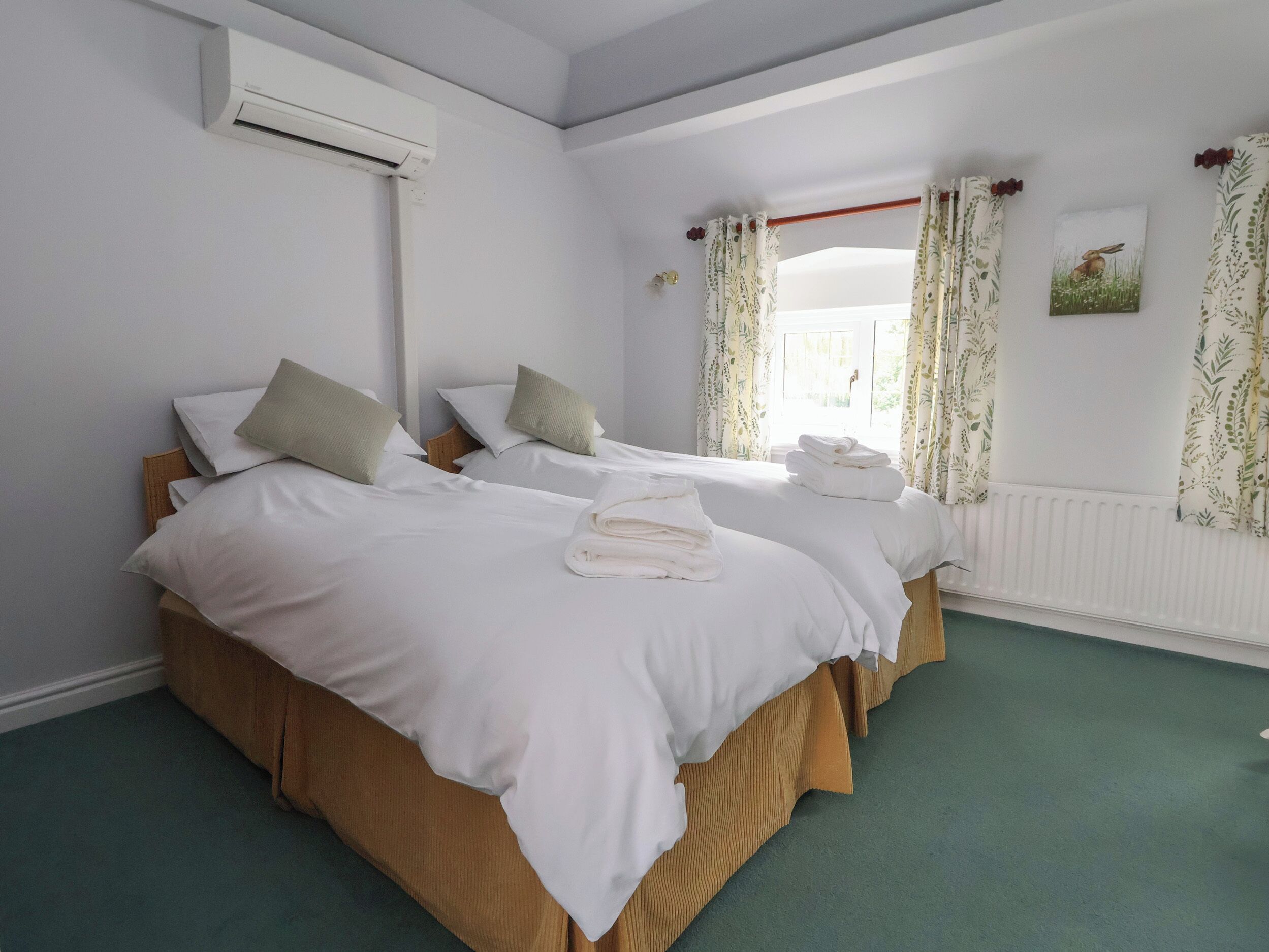 Une chambre d’hôtel avec deux lits, du linge de lit blanc, un radiateur et une fenêtre agrémentée de rideaux à motifs floraux.