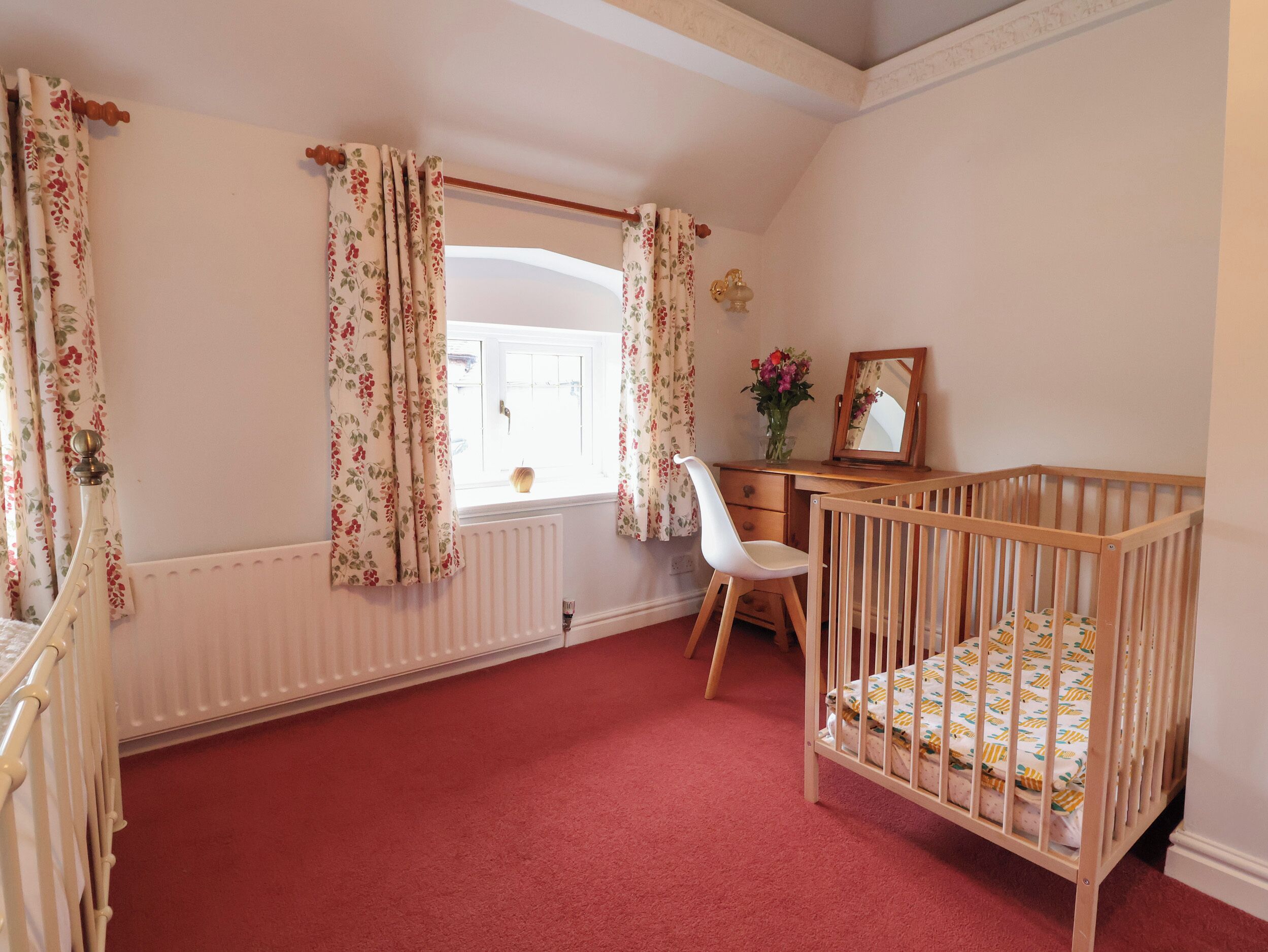 Ein Babyzimmer mit einem Kinderbett, einem Schreibtisch, einem Stuhl, einer Heizung und blumigen Vorhängen.