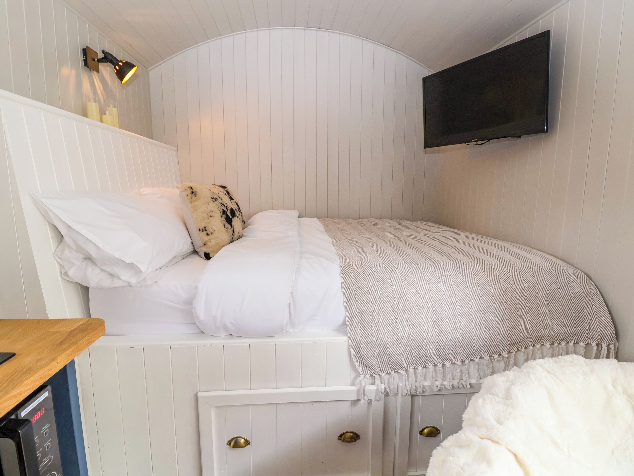 A neatly arranged bedroom with a bed, a mounted television, a wall-mounted lamp, and a small wooden table.