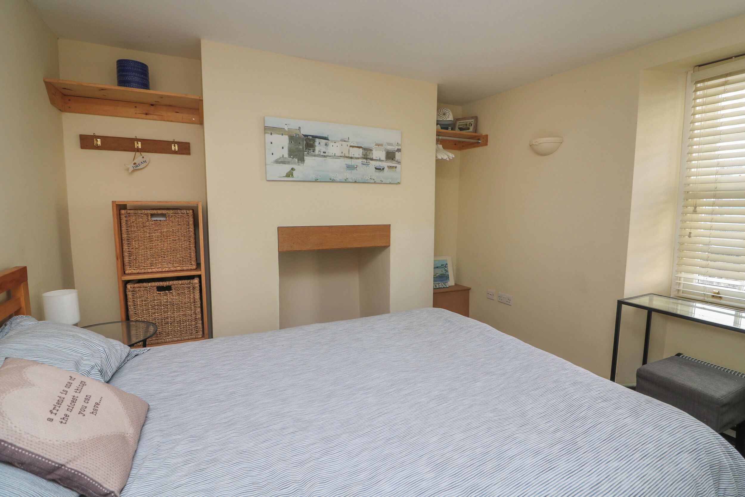 A bedroom with a bed, a small table, a chair, a window with blinds, and wall-mounted shelves with various items.