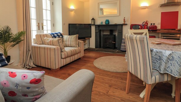 A cozy living room with a sofa, armchair, and a dining table with chairs. There's a fireplace and a television. - Quarter Deck (Salcombe)
