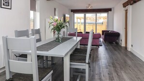 Un coin repas avec une table blanche, des chaises grises et un vase rempli de fleurs. On peut y admirer une poutre en bois au plafond et, à l’extérieur, une terrasse en bois.