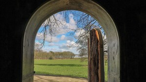 Una porta en arc que dona a una zona empavedada de pedra amb vistes a un camp verd i arbres.