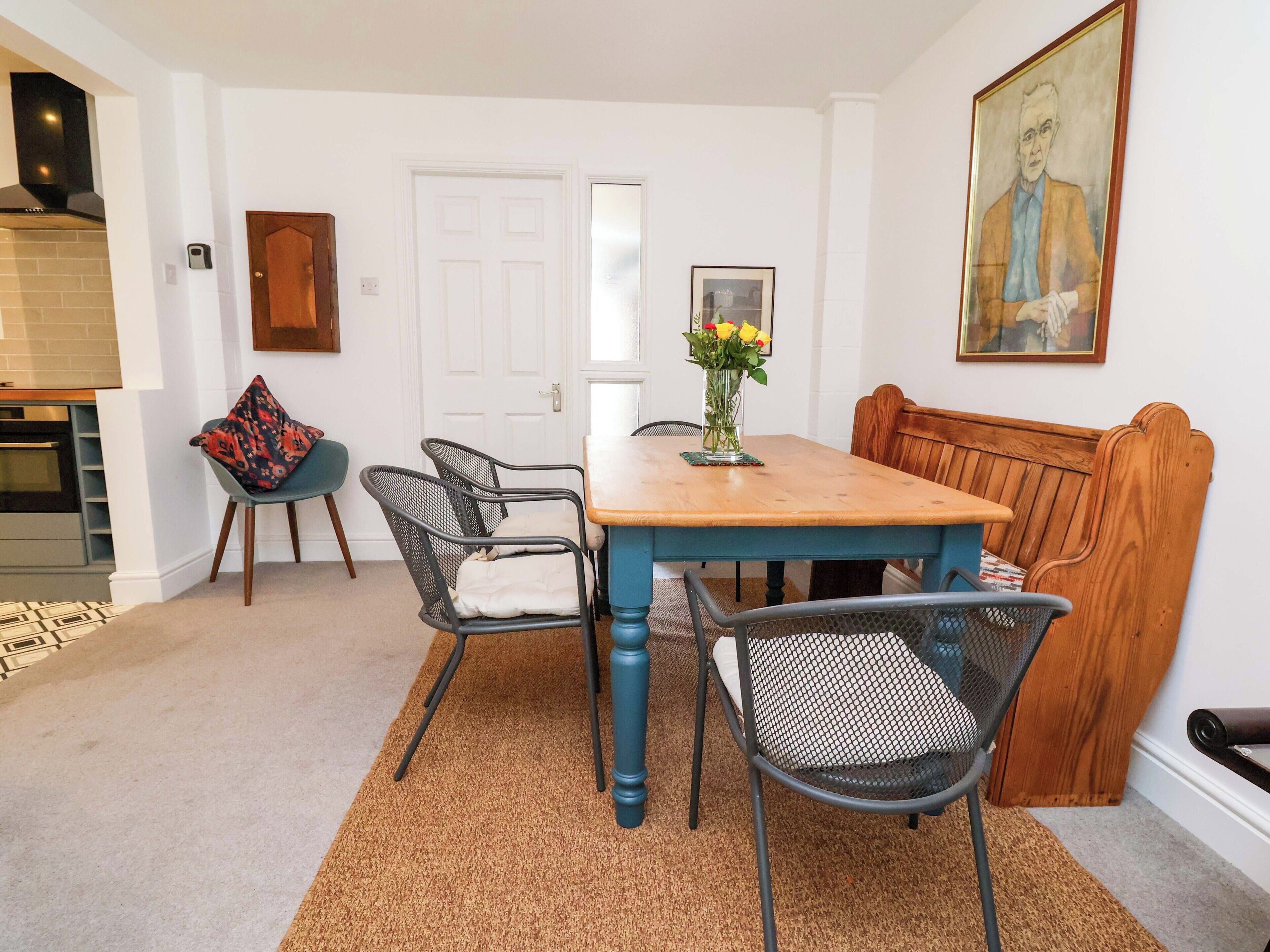 Un comedor con una mesa y sillas de madera, un jarrón con flores, un cuadro en la pared y una puerta.